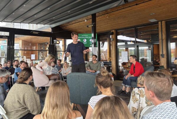 A man stands in the center of a conference room, speaking to people sat on chairs around him